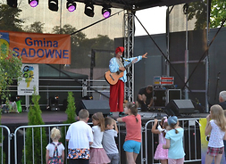 Na scenie stoi mężczyzna w czapce krasnoludka i wskazuje w lewo lewą ręką. Prawą ręką przytrzymuje gitarę. Przed sceną przy barierkach widać grupkę dzieci