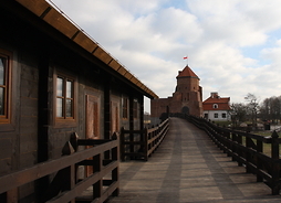 Po lewej stronie widać drewniany niski budynek z barierką. Obok budynku biegnie drewniana droga - pomost, który prowadzi do zamku z flagą Polski na szczycie
