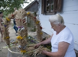 Kobieta przed drewnanym domem uplata dożynkowy weniec z suszonych kwiatów i kłosów zbóż