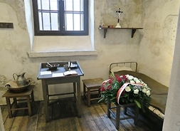 Wnętrze celi Traugutta ze skromnymi sprzętami: drewnianym stolikiem, dwoma taboretami, metalowym łóżkiem, krzyżem stojącym na półcenad nad posłaniem. Pośrodku zakratowane okno celi.