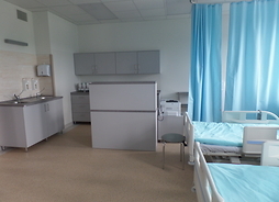Po prawej stronie - widać trochę dwa łóżka szpitalne przedzielone kotarą, po lewej - część sanitarna sali, z szafkami i umywalką