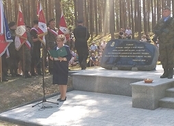 Elżbieta Lanc stoi przy mikrofonie i przemawia. Za nią jest kamienny szeroki pomnik, który tworzy przepołowiony na pół głaz z napisaną sentencją. Po lewej stronie widać flagi i kombatantów