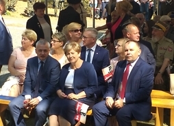Grupa ludzi siedzi na krzesłach. Są zwróceni twarzą do kamery. Na pierwszym planie siedzi Elżbieta Lanc, obok niej kombatanci. Wszyscy trzymają flagi Polski z symbolem Polski walczącej