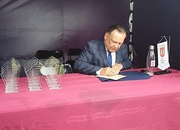 Marszałek Adam Struzik siedzi pod namiotem przy stole. Podpisuje dokument. Po jego prawej stronie stoją przezroczyste statuetki z wypisanym podziękowaniem