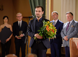 Janusz Wawrowski stoi na scenie, trzyma w ręku kwiaty, za nim stoją pozostali laureaci nagród norwidowskich