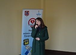 Janina Ewa Orzełowska stoi, w prawym ręku trzyma mikrofon, do którego mówi. Za nią jest baner instytucji