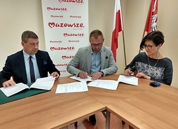 Dwóch mężczyzn i kobieta siedzą przy stole i podpisują dokumenty. Za nimi jest baner z logiem Mazowsze oraz flagi Polski i województwa