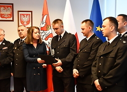 Członek zarządu Janina Ewa Orzełowska przekazuje teczkę mężczyźnie w srażackim garniturze. Po ich obu stronach stoją inni przedstawciele OSP