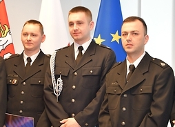 Trzech młodych mężczyzn w garniturach strażackich stoi na tle flag UE, Polski i województwa