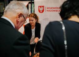 Przed stoiskiem promocyjnym Mazowsza dwie osoby zwiedzające stoją tyłem do obiektywu, na stoisku kobieta prezentuje materiały informacyjne.