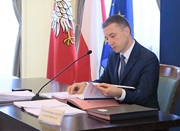 Przewodniczący Sejmiku Województwa Mazowieckiego Ludwik Rakowski siedzi za stołem prezydialnym i odczytuje przeglądane dokumenty.