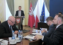Trzech radnych klubu Koalicja Obywatelska siedzi za stołem obrad, za nimi przemawia z trybuny Dariusz Grajda, członek zarządu i dyrektor handlowy Kolei Mazowieckich.