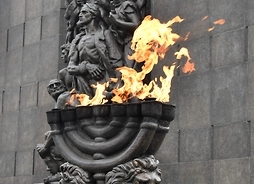 Wieczny ogień pamięci, płonący na tle Pomnika Bohaterów Getta w Warszawie.