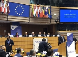 Widok na główną ławę prowadzących zebranie komitetu. Przed nią jest ambona, w której stoi mężczyzna w garniturze. U góry nad ich głowami wisi flaga UE oraz telebim z nazwiskiem przemawiającego