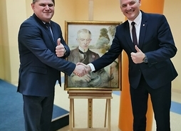 Wicemarszałek i dyrektor Ruszczyk podają sobie prawe dłonie na tle portretu Podkańskiego. Lewe dłonie mają uniesione z podniesionym kciukiem do góry