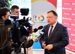 Marszałek Adam Struzik udziela wywiadu telewizyjnegostojąc przodek do kamery i dwóch dziennikarzy, na tle roll-upu z emblematem BOM i śnianki z logotypem Marki Mazowsze.