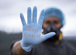 Sylwetka medyka w stroju ochronnym z rozpostartą dłonią w rękawiczce hirurgicznej niebieskiej z czerwonym napisem "Stop Covid-19"