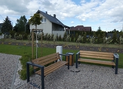 Fragment zagospodarowanej strefy rekreacji: dwie ławki ogrodowe na wubrukowanych kostką placyku, a tyłu w tle stoi dom mieszkalny jednorodzinny.