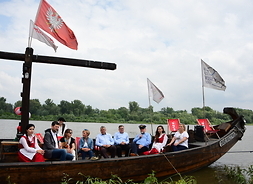 Na rzece. w drewnianej wielkiej łodzi siedzą marszałek Struzik, dyrektor Stelmańska i dyrektorzy muzeów. Obok nich są dwie dziewczyny w ludowych strojach i trzej grajkowie. Marszałek trzyma w ręku mikrofon