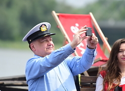 Marszałek w czapce kapitana trzyma w wyciągniętych rękach aparat. Zdjęcie z boku, od pasa w górę. W tle widać wodę i drzewa z przeciwegłego brzegu rzeki