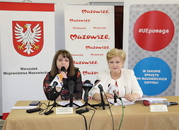 Dwie kobiety siedzą za stołem prezydialnym, przed mikrofonami. Siedząca z lewej mówi do mikrofonu.