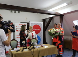 Dwóch ratowników medycznych przekazuje kwiaty dwóm kobietom stojacym za stołem prezydialnym. Z lewej kamerzysta nagrywa przekaz..