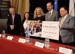 przedstawiciele samorządu Mazowsza i Przasnysza stoją na sali konferencyjnej obok siebie, trzymając symboliczny czek w ręku