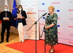 Elżbieta Lanc stoi i mówi do mikrofonu, obok w oddaleniu stoją kobieta i męzczyzna w maseczkach.