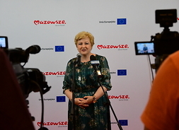 Elżbieta Lanc mówi do mikrofonów dziennikarskich, jest nagrywana przez dwie kamery telewizyjne.