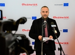 Tomasz Rosłonek stoi i mówi do mikrofonu, filmowany przez kamerę telewizyjną.y