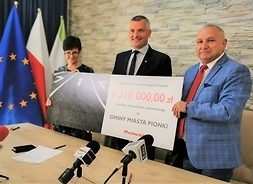 Wicemarszałek Rafał Rajkowski wraz z burmistrzem i skarbniczką stojąc trzymają plakat tekturowy informujacy o inwestycji.