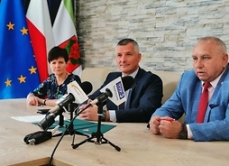 Wicemarszałek Rafał Rajkowski wraz z burmistrzem i skarbniczką siedzą za stołem z rozstawionymi mikrofonami.