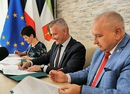 Wicemarszałek Rafał Rajkowski wraz z burmistrzem i skarbniczką siedzą za stołem i podpisują dokumenty.