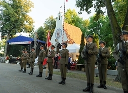 W dużym planie widoku z prawej strony od frontu duża, udekorowana scena , na jej tle stoi w rzędzie ośmiu żołnierzy umundurowanych.