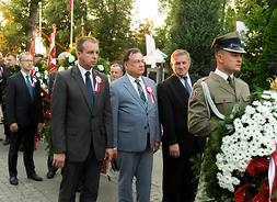 Marszałek Adam Struzik wraz z dwoma innymi członkami delegacji, podczas podejścia do składania wieńca, który niesie przed nim żołnierz w mundurze, w tle kilku innych uczestników.