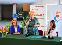 Trzy kobiety za stołem prezydialnym, dwie po bokach siedzą, ta w środku stoi i mówi do mikrofonu.