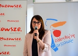 Na tle rollupów z logotypami Mazowsza i Warszawy stoi kobieta w okularach i mówi do mikrofonu.