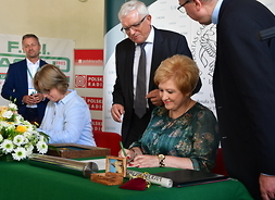 Elżbieta Lanc przy stole podpisuje dokument. Obok niej siedzi kobieta pochylona nad innym dokumentem. Za Elżbietą Lanc stoją lekko pochyleni dwaj mężczyźni w garniturach