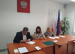 Trzy osoby siedzą za stołem podpisując dokumenty.