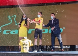 Na scenie mężczyzna w kolarskim stroju trzyma w rękach otwarty szampan, który tryska do góry. Po jego prawej stronie stoi długowłosa dziewczyna, a po lewej - mężczyzna w garniturze. Wszyscy mają maski na twarzy