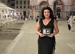 Alicja Węgorzewska stoi na tle zabytkowego monumentalnego budynku we Włoszech. W rękach przed sobą trzyma nagrodę.