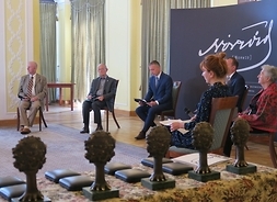 Siedmioro uczestników siedzi na osobnych krzesłach w kręgu, na pierwszym planie stół ze statuetkami nagrody Norwida.