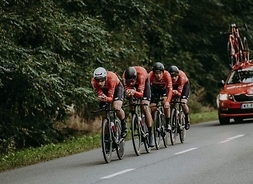 Czterech zawodników na rowerach wyścigowych jedzie na szosie jeden za drugim, za nimi jedzie wóz techniczny z zapasowymi rowerami na dachu.