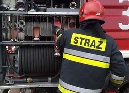 Tyłem stoi strażak z OSM w umundurowaniu bojowym na tle samochodu ratowniczo-gaśniczego.