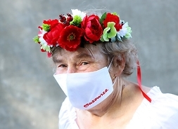 Kobieta w dopasowanej maseczne białej z czerwonym logo samorządu Mazowsza, na głowie wianek ze swieżych kwiatów.