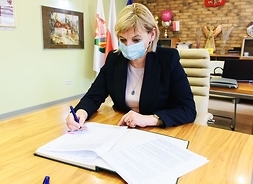 Kobieta w maseczce siedzi za stołem i podpisuje dokumenty.