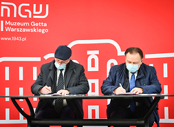 Dwóch męzczyzn w maseczkach siedzi z stołem i podpisuje dokumenty. Za nimi banner z logotypem Muzeum Getta Warszawskiego.