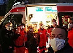 Osiem osób w rzędzie i w grupie, twarze w maseczkach ochronnych, za nimi ambulans medyczny z odsłoniętym wnętrzem.