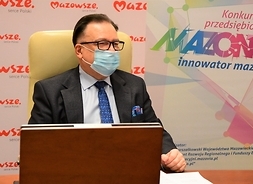 Marszałek siedzi w maseczce ochronnej przed ekranem komutera, za nim bannerry zz logotypem Mazowszai z logotypem konkursu.