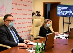 Dwie osoby w maseczkach ochronnych siedzą za stołem przed ekranami komputerów podczas spotkania, z tyłu za nimi baner z logotypem samorządu Mazowsza, po prawej u góry duży ekran pokazujący transmisję ze spotkania.